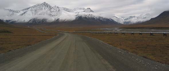 Dalton Highway approaching snow-capped Brooks Range in distance, with pipeline along right side of the road